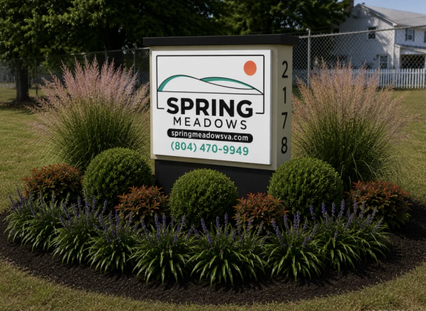 A rectangular outdoor sign for Spring Meadows with contact info, surrounded by neatly trimmed bushes, flowers, and mulch. A house and chain-link fence are visible in the background.
