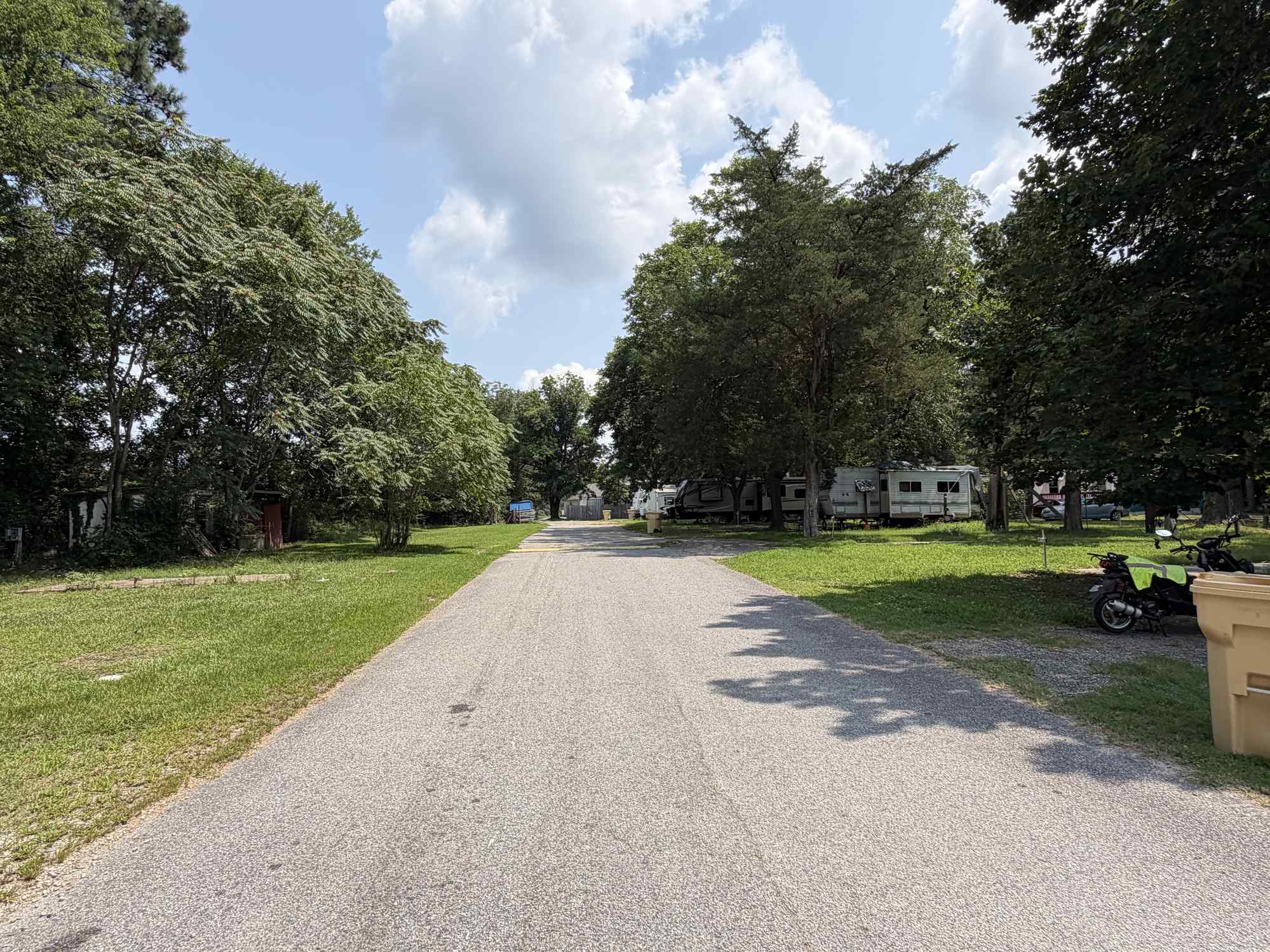 A paved road runs between grassy areas with trees on both sides; RVs and a motorcycle are visible in the background under a partly cloudy sky.