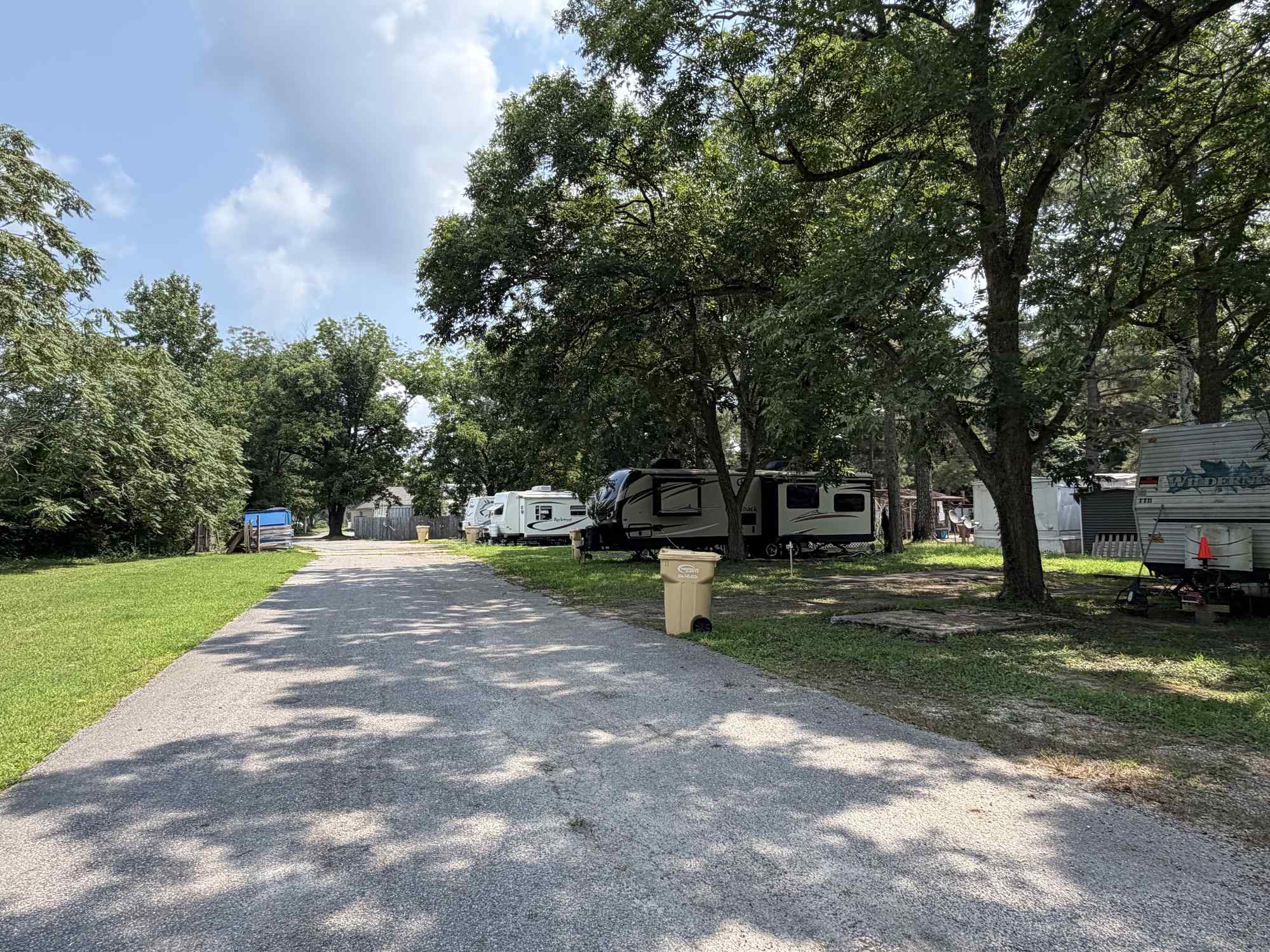 Paved road in a campground with RVs parked under trees on the right side, grassy area on the left, and a trash bin near the road.