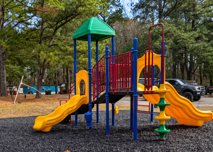 Colorful playground equipment with slides and climbing structures stands on a rubber surface, surrounded by trees and parked vehicles in a residential outdoor setting.